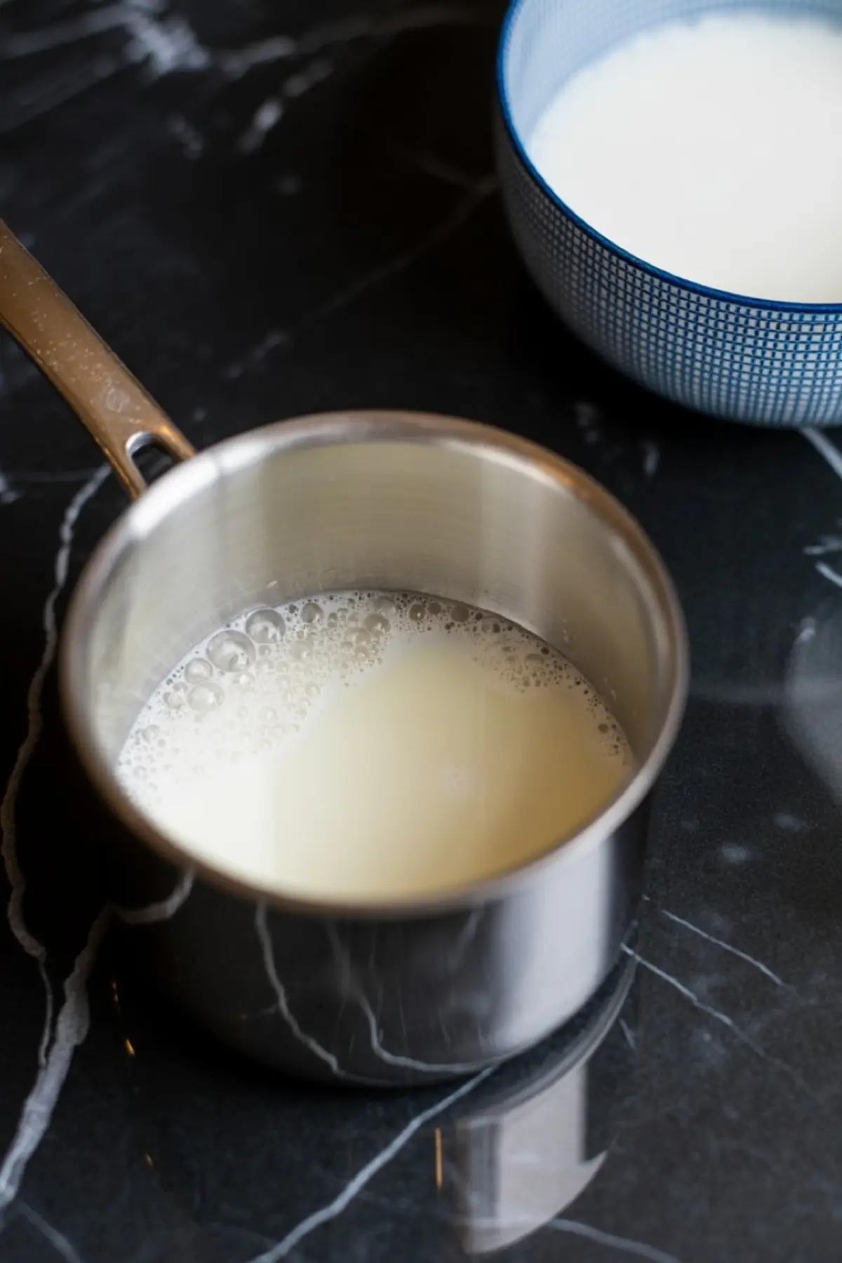 Stainless steel saucepan filled with warm milk sits on a black marble countertop, showing the early step of a homemade caramel coffee recipe.
