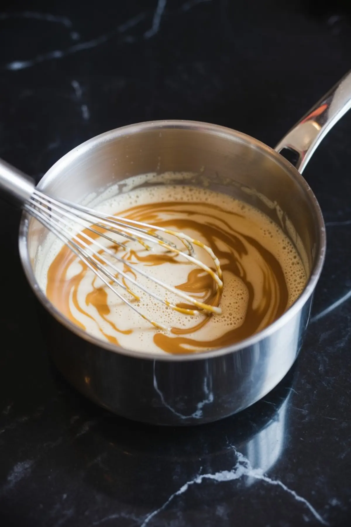 Caramel coffee mixture swirled with golden caramel sauce in a saucepan being whisked on a black marble countertop, showing the blending process.
