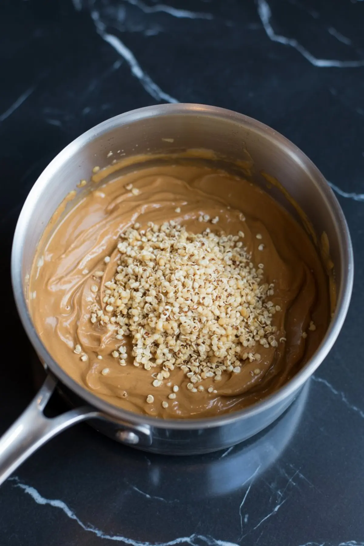 Cooked quinoa added to a smooth peanut butter mixture in a saucepan, showing the mixing stage for peanut butter quinoa energy bites on a dark countertop.