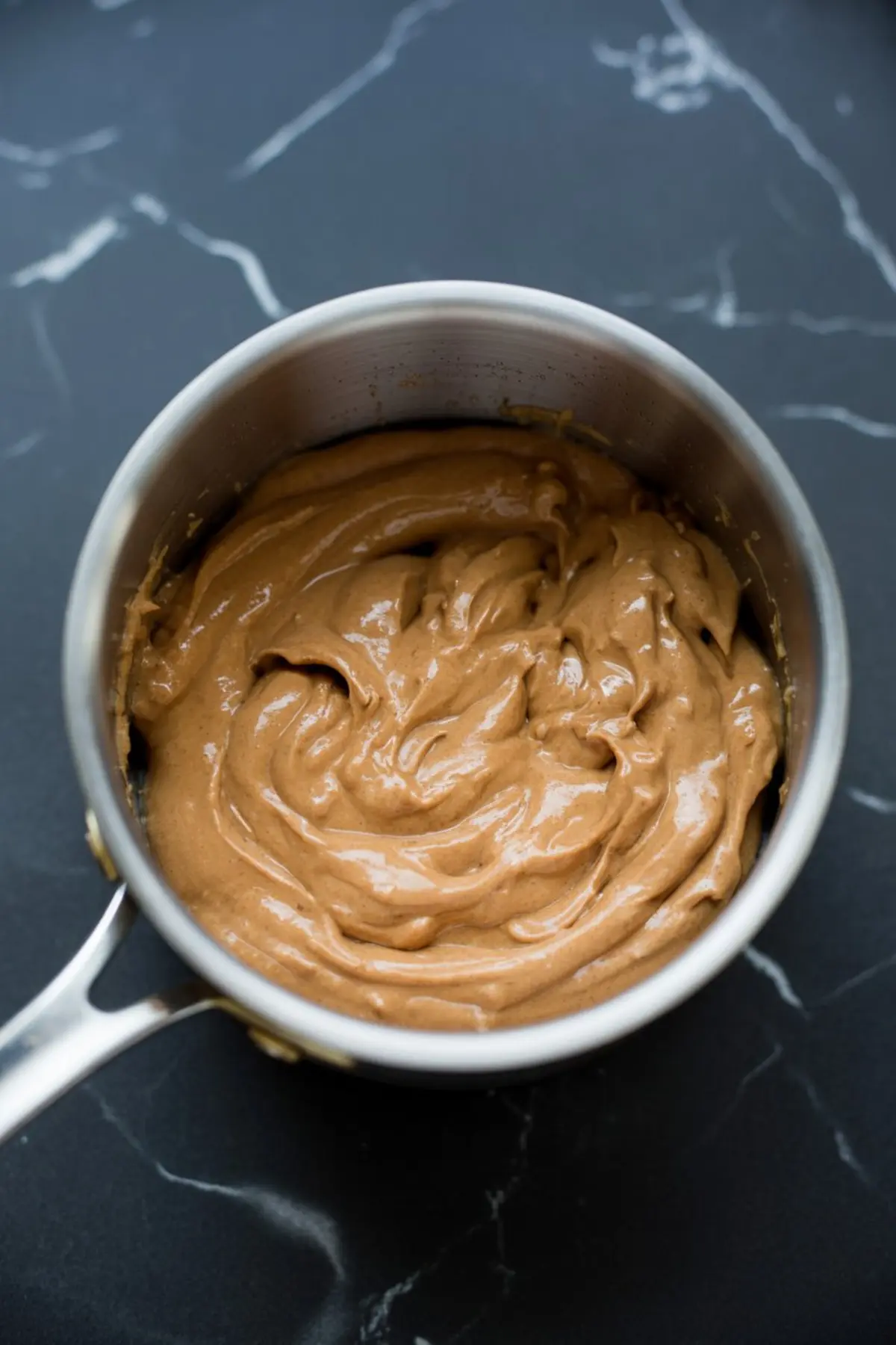 Smooth peanut butter mixture swirled inside a metal saucepan on a dark marble countertop, prepared for making peanut butter quinoa snack bites.