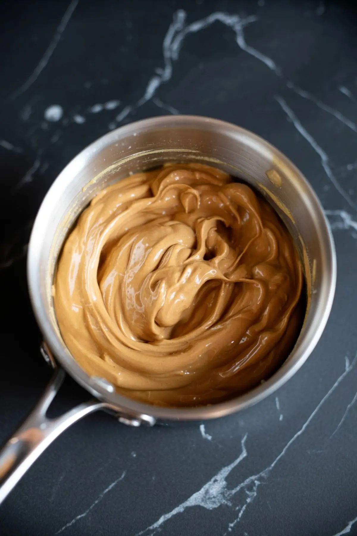 Creamy peanut butter blended in a saucepan with a thick glossy texture, photographed from above on a dark stone surface for a no bake snack recipe.