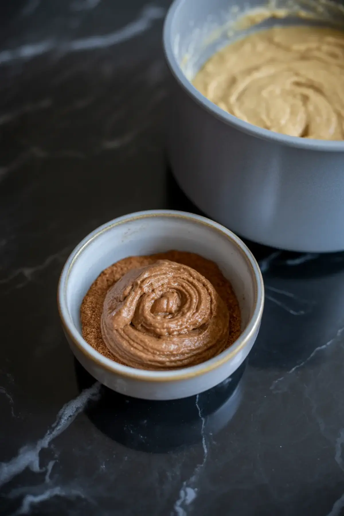 Small bowl filled with cinnamon sugar topped by a swirl of cinnamon filling, with a large bowl of thick muffin batter in the background on a dark marble surface.