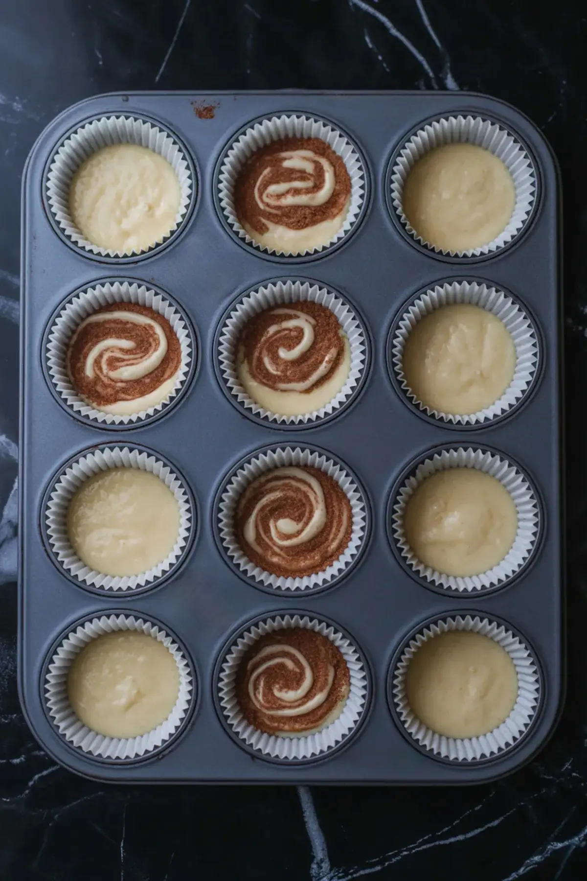 Muffin tray filled with raw sour cream muffin batter, some cups featuring a visible swirl of cinnamon sugar mixture, ready for baking on a dark marble counter.