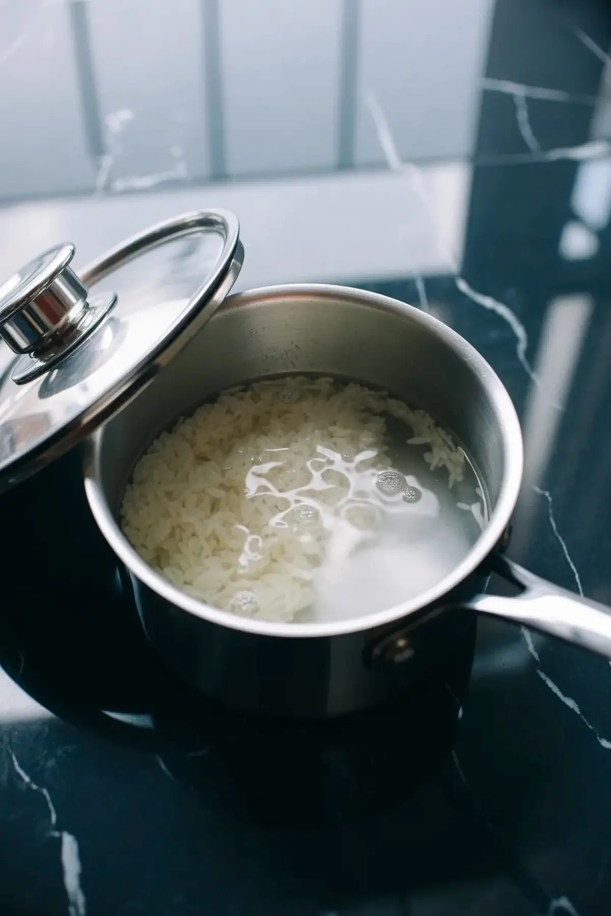 A metal saucepan holds white rice simmering in water on a stovetop. The pot lid rests slightly open, and steam forms small bubbles on the surface. This cooking process image shows rice boiling for a homemade rice pudding recipe.
