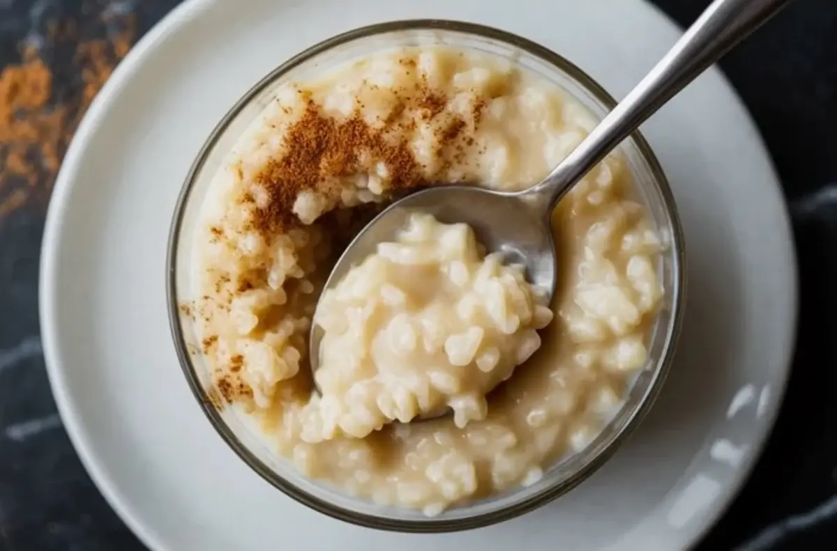 A spoon lifts creamy rice pudding from a glass bowl topped with ground cinnamon. The pudding sits on a white plate, and the texture shows soft rice grains in thick custard. This close up dessert image highlights homemade rice pudding with condensed milk and warm cinnamon spice.

