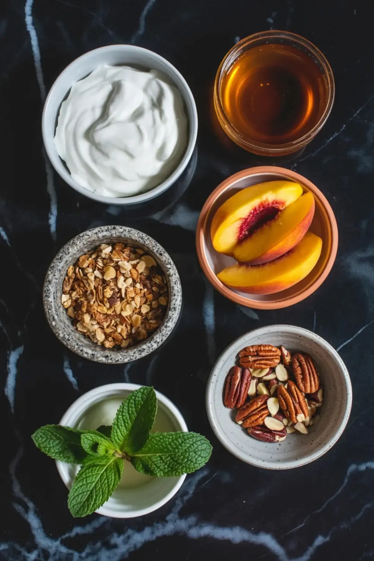 Ingredients for peach yogurt bark are arranged in bowls with Greek yogurt, honey, peach slices, granola, pecans, and fresh mint on a dark marble surface.