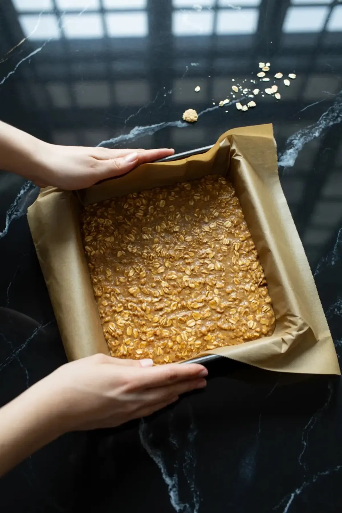 The oat base for blueberry oatmeal bars is pressed firmly into a parchment-lined square pan before the filling is added.