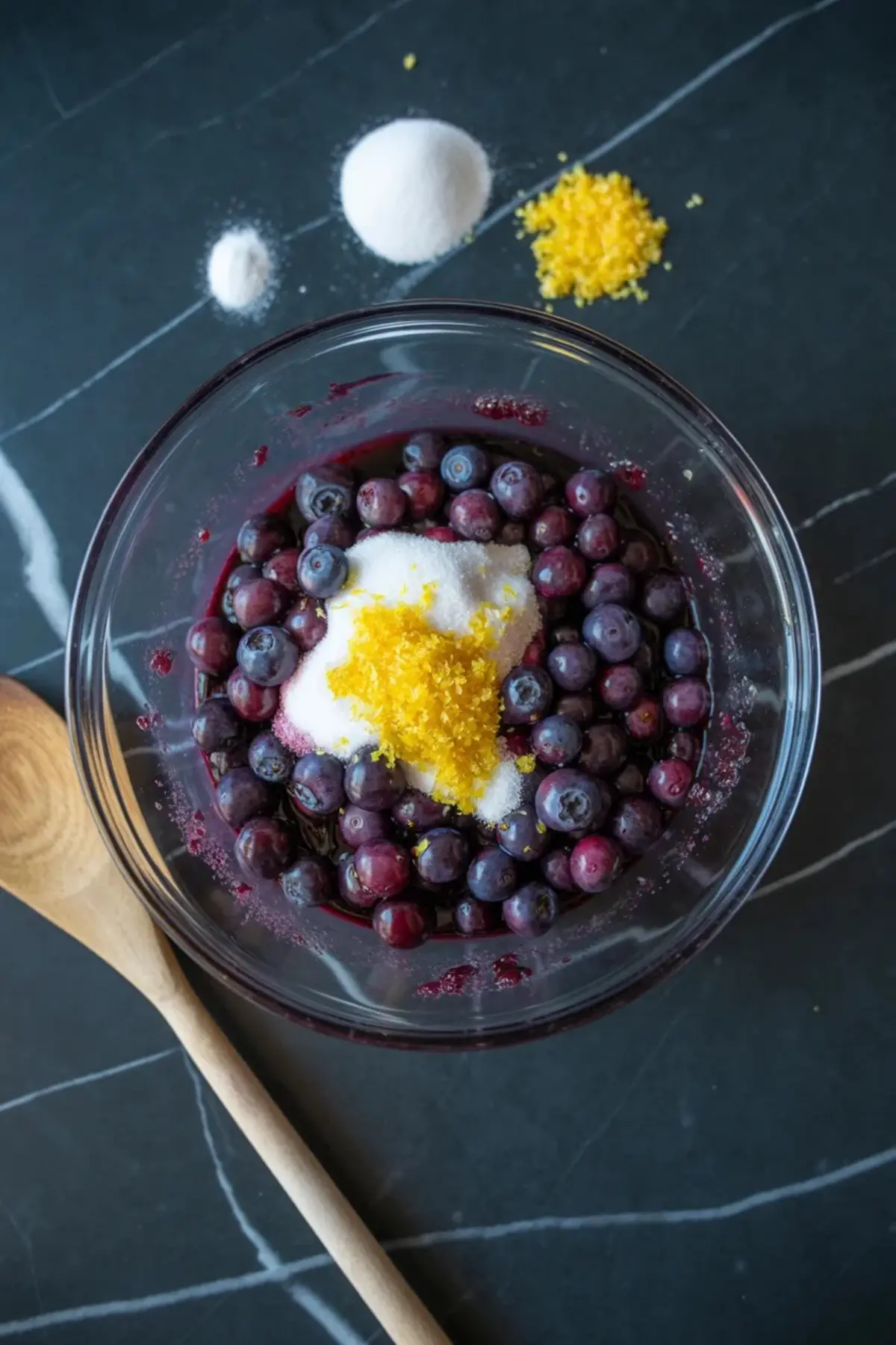 Blueberries are mixed with sugar and lemon zest in a glass bowl while the filling for blueberry oatmeal bars is prepared.