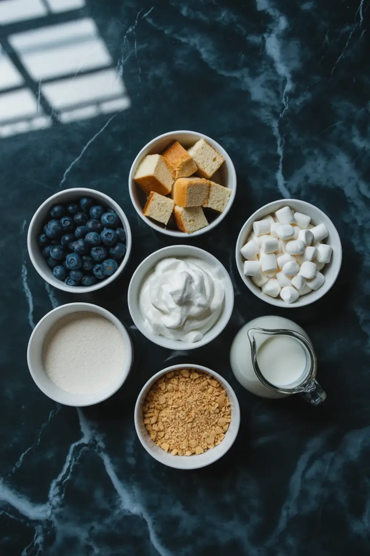 Ingredients for blueberry fluff salad are arranged with blueberries, cake cubes, mini marshmallows, whipped topping, milk, sugar, and crumbs on a dark surface.