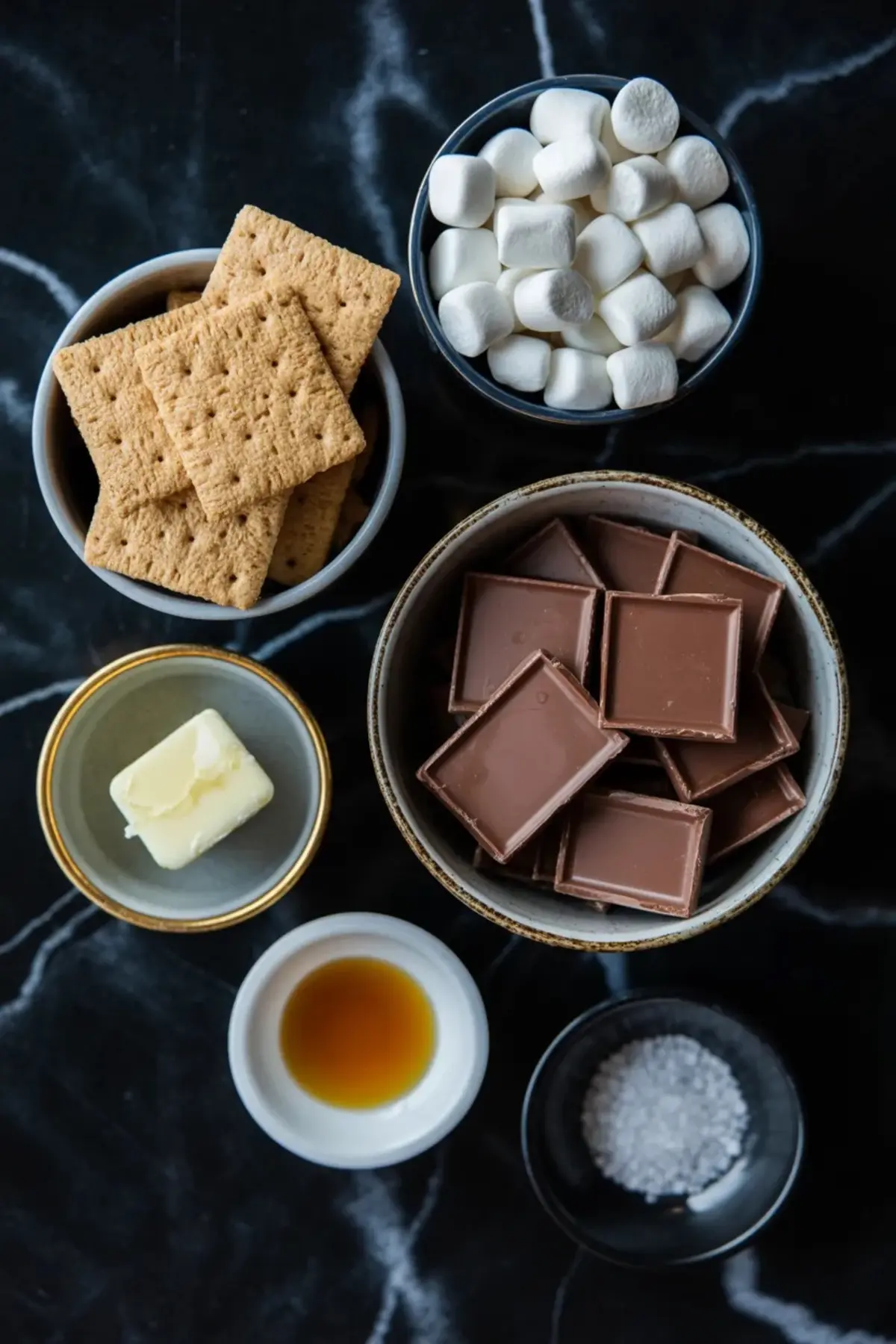 Ingredients for Golden Grahams s’mores bars are arranged with graham crackers, mini marshmallows, milk chocolate, butter, vanilla, and salt on a dark surface.