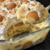 A spoonful of old-fashioned banana pudding lifted from a glass 9x13 dish, showing vanilla custard, a banana slice and a soft whole vanilla wafer under whipped cream, with custard and wafer layers visible through the side of the dish on a black stone counter.