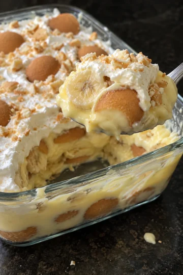 A spoonful of old-fashioned banana pudding lifted from a glass 9x13 dish, showing vanilla custard, a banana slice and a soft whole vanilla wafer under whipped cream, with custard and wafer layers visible through the side of the dish on a black stone counter.