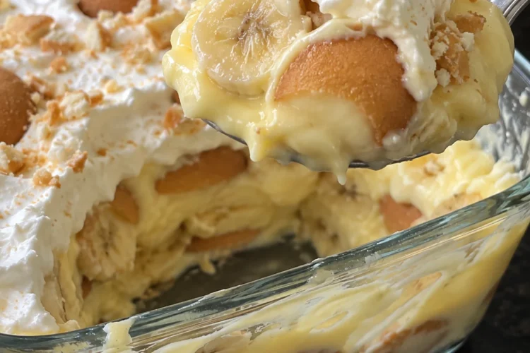 A spoonful of old-fashioned banana pudding lifted from a glass 9x13 dish, showing vanilla custard, a banana slice and a soft whole vanilla wafer under whipped cream, with custard and wafer layers visible through the side of the dish on a black stone counter.