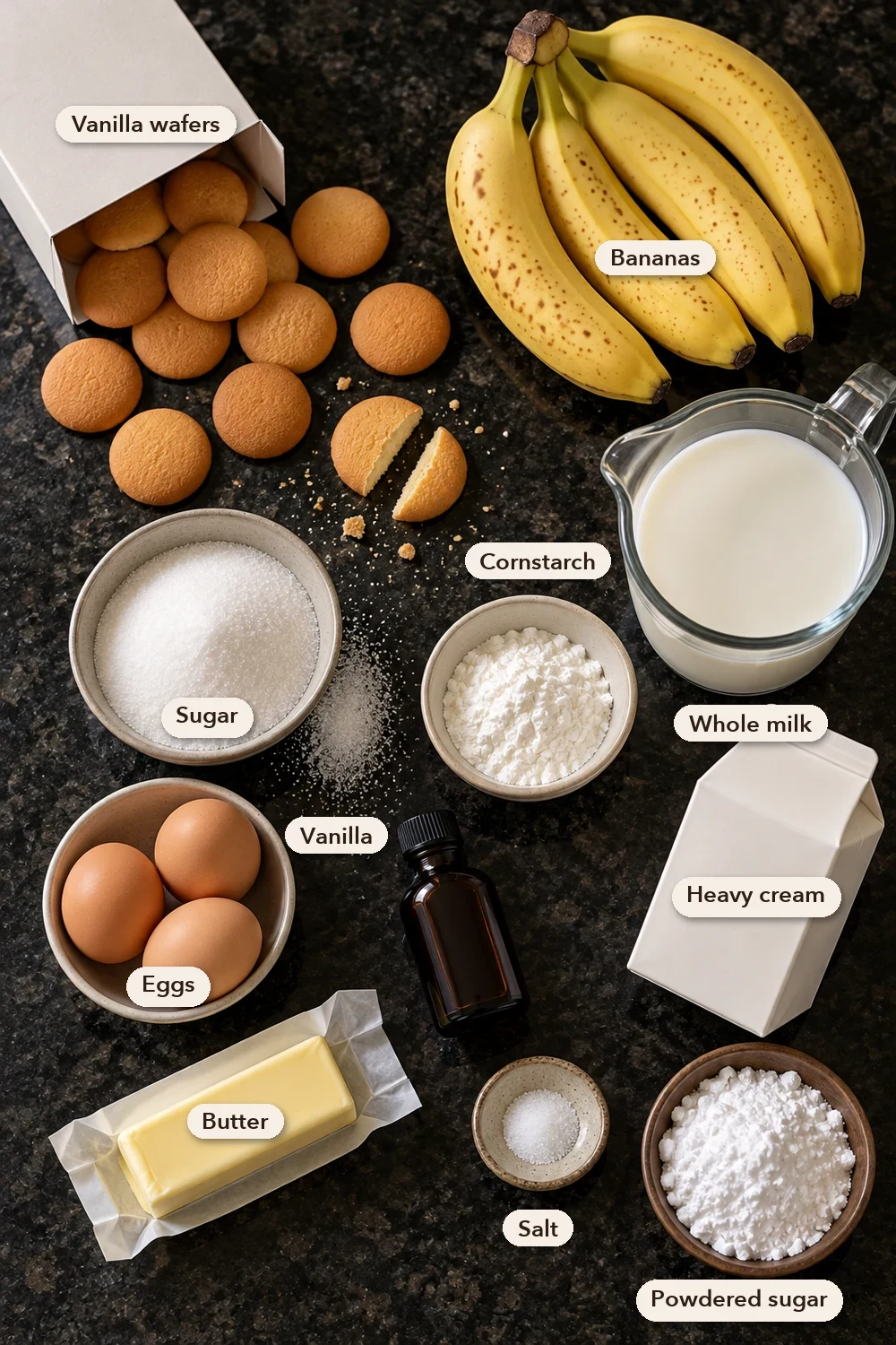 Labeled ingredients for homemade banana pudding: vanilla wafers, ripe bananas, whole milk, sugar, cornstarch, eggs, vanilla, heavy cream, butter, salt and powdered sugar on a black stone counter.