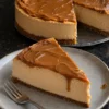 A slice of no-bake Biscoff cheesecake on a grey stoneware plate with a fork, a glossy cookie butter top dripping down the cut side over a pale caramel filling and a thin cookie crust, the rest of the cheesecake behind it with speculoos cookies standing in the top.
