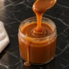 A teaspoon lifting a thick ribbon of glossy amber caramel sauce over a small glass jar, a drip running down the jar into a little puddle on a black marble counter.