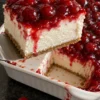 A square of cherry delight dessert lifted from a white 9x13 dish on an offset spatula, with glossy cherry pie filling dripping down a thick white cream cheese layer over a thin graham crust, on a black stone counter.