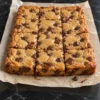 Twelve chocolate chip cookie bars cut in the slab on crinkled parchment on a black marble counter, with golden crackled tops, glossy pools of melted chocolate and one loose chip on the paper.