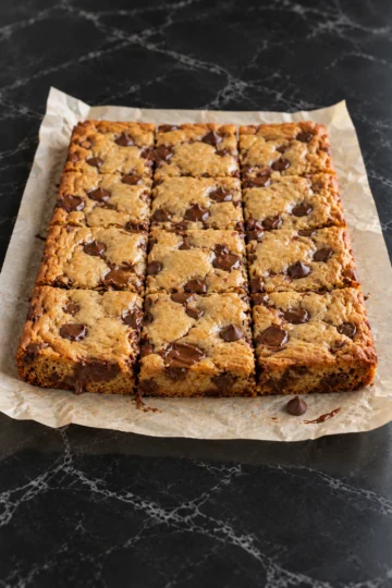 Twelve chocolate chip cookie bars cut in the slab on crinkled parchment on a black marble counter, with golden crackled tops, glossy pools of melted chocolate and one loose chip on the paper.