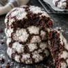 A stack of 4 chocolate crinkle cookies on a black stone counter, the top one broken open to show a dark, fudgy center under cracked white powdered sugar.