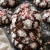 A stack of 4 chocolate peppermint crinkle cookies on a black stone counter, with dark fudgy cracks, thick white powdered sugar and crushed red and white candy cane pressed into the top.