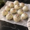White chocolate Oreo balls on crinkled parchment on black marble, topped with a random scatter of red and green nonpareils, a few with a white drizzle, a pine sprig at the edge.