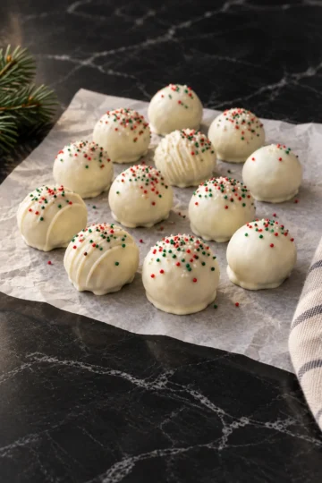 White chocolate Oreo balls on crinkled parchment on black marble, topped with a random scatter of red and green nonpareils, a few with a white drizzle, a pine sprig at the edge.
