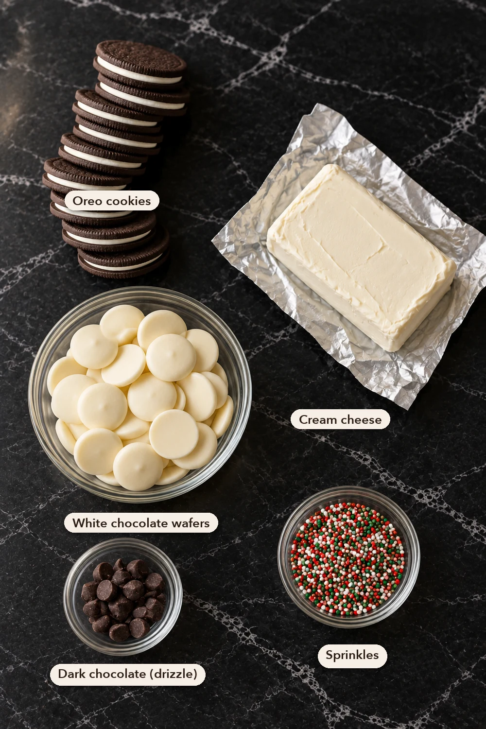 Labeled Oreo ball ingredients from above on black marble: a stack of Oreo cookies, a block of cream cheese on its foil, a bowl of white chocolate wafers, a dish of dark chocolate chips and a dish of red and green sprinkles.