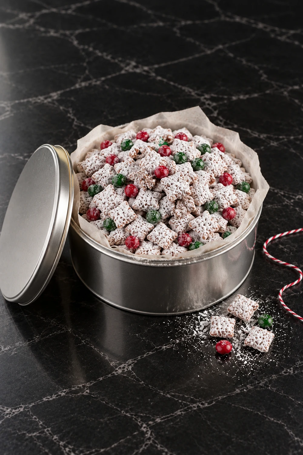 A silver cookie tin lined with parchment and filled to the brim with Christmas puppy chow, its lid leaning against the side and a loop of red and white baker's twine on the marble.