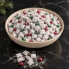 A cream stoneware bowl heaped with Christmas puppy chow, small cereal squares in a thick white powdered sugar coat mixed with red and green candy-coated chocolates, a few pieces and a drift of sugar spilled on black marble and a pine sprig at the edge.