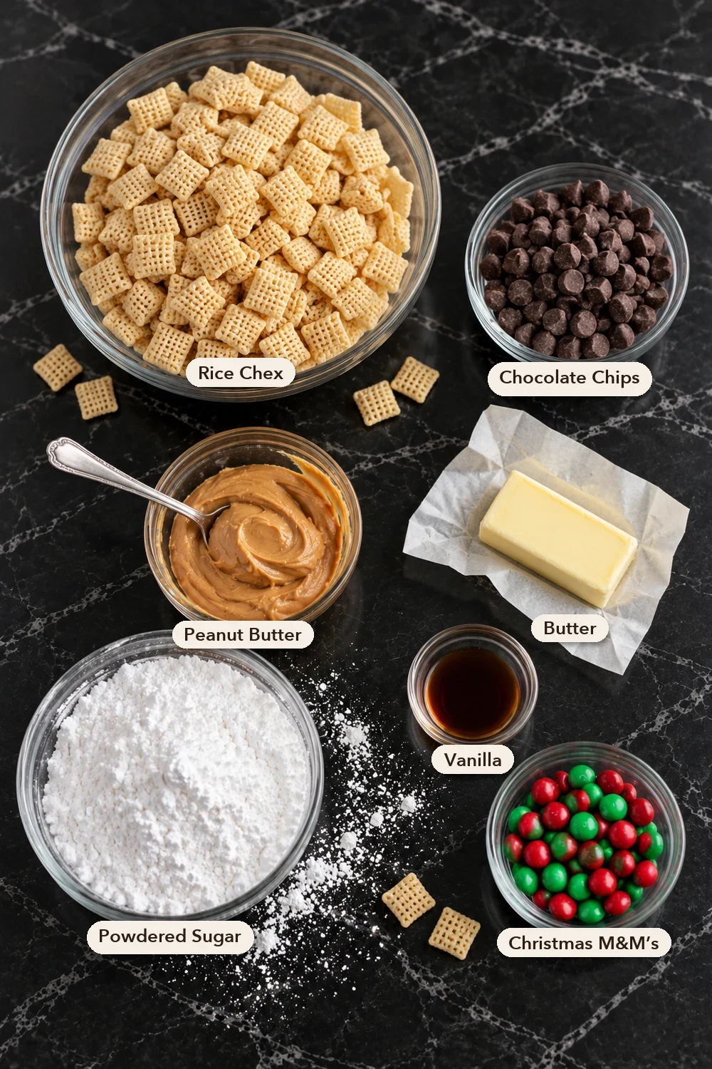 Labeled puppy chow ingredients seen from above on black marble: a large glass bowl of Rice Chex, chocolate chips, peanut butter with a spoon, a half stick of butter on its wrapper, vanilla, a bowl of powdered sugar and red and green M&Ms.