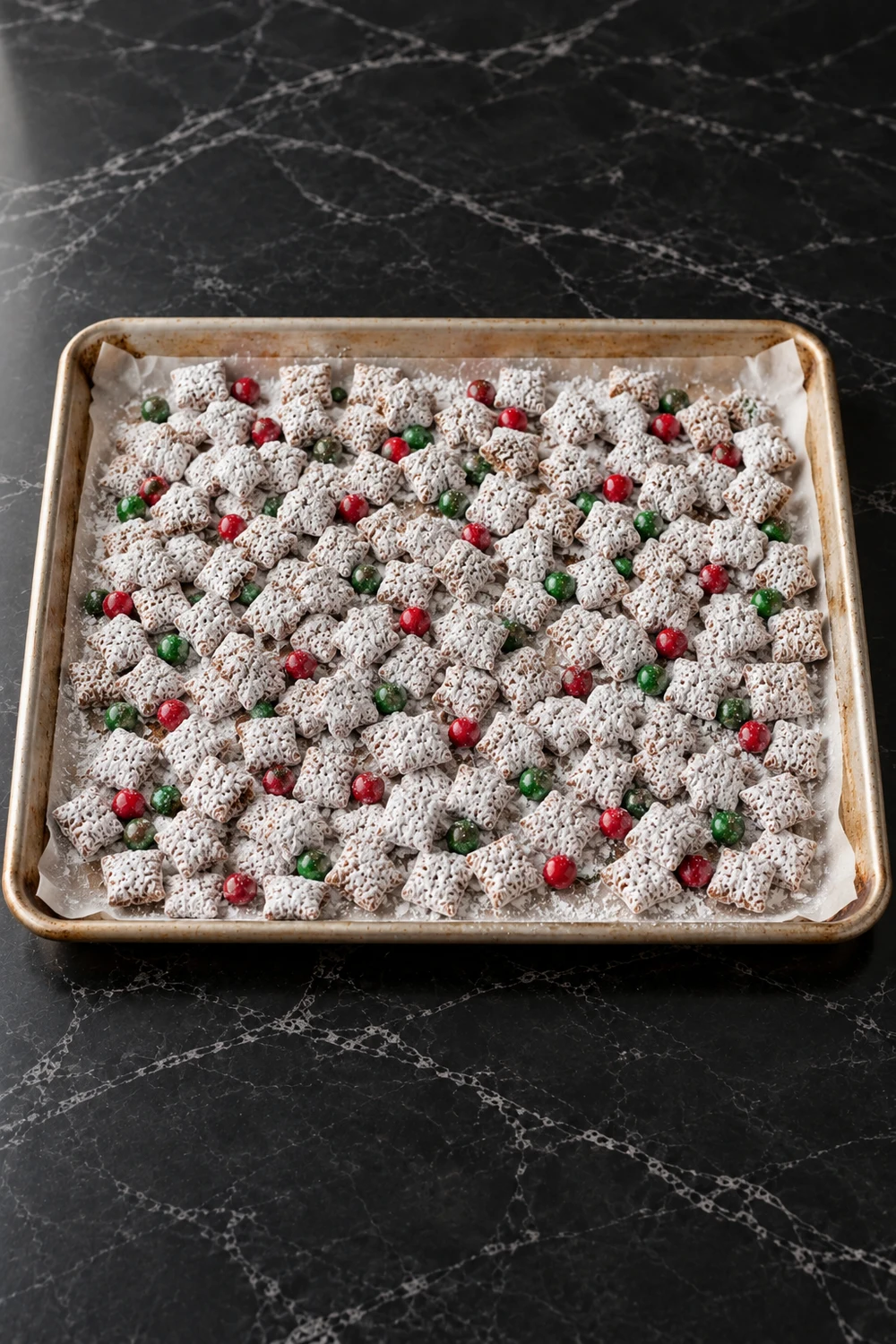 Sugar-coated puppy chow spread in one layer on a parchment-lined sheet pan to set, with red and green M&Ms scattered over it and powdered sugar dusting the paper.