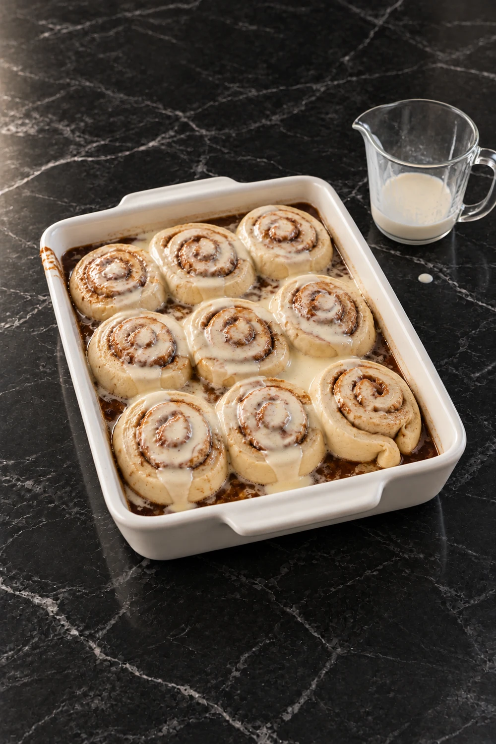 Ten raw cinnamon rolls sitting on butter and brown sugar in a white 9x13 dish, with heavy cream just poured over them and pooling in the gaps, and a glass measuring cup beside it.