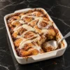 A white 9x13 dish of cinnamon roll dump cake with golden swirled rolls, ribbons of cream cheese icing and dark caramel sauce bubbling at the edges, one corner scooped out with a serving spoon resting in the gap.