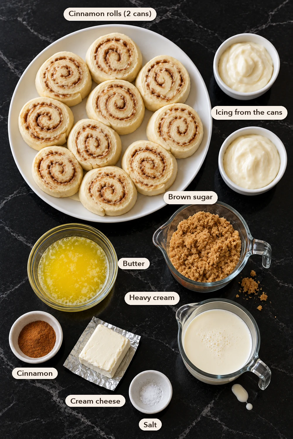 Labeled ingredients on black marble: a plate of ten raw canned cinnamon rolls, two cups of icing, melted butter, brown sugar, heavy cream, ground cinnamon, a small block of cream cheese and salt.
