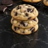A stack of three thick cookies and cream cookies studded with Oreo chunks and white chocolate chips on a black marble counter, with two Oreos beside them and more cookies on a cooling rack behind.