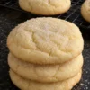 A stack of three thick cream cheese cookies with pale cracked tops and a sparkly sugar coat on a black stone counter, more cookies cooling on a wire rack behind them.