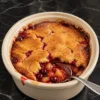 A cream stoneware slow cooker crock of cherry pineapple dump cake with a cracked golden buttery topping and glossy red cherry filling bubbling through the gaps, one spoonful scooped out at the front with a serving spoon resting in the fruit.