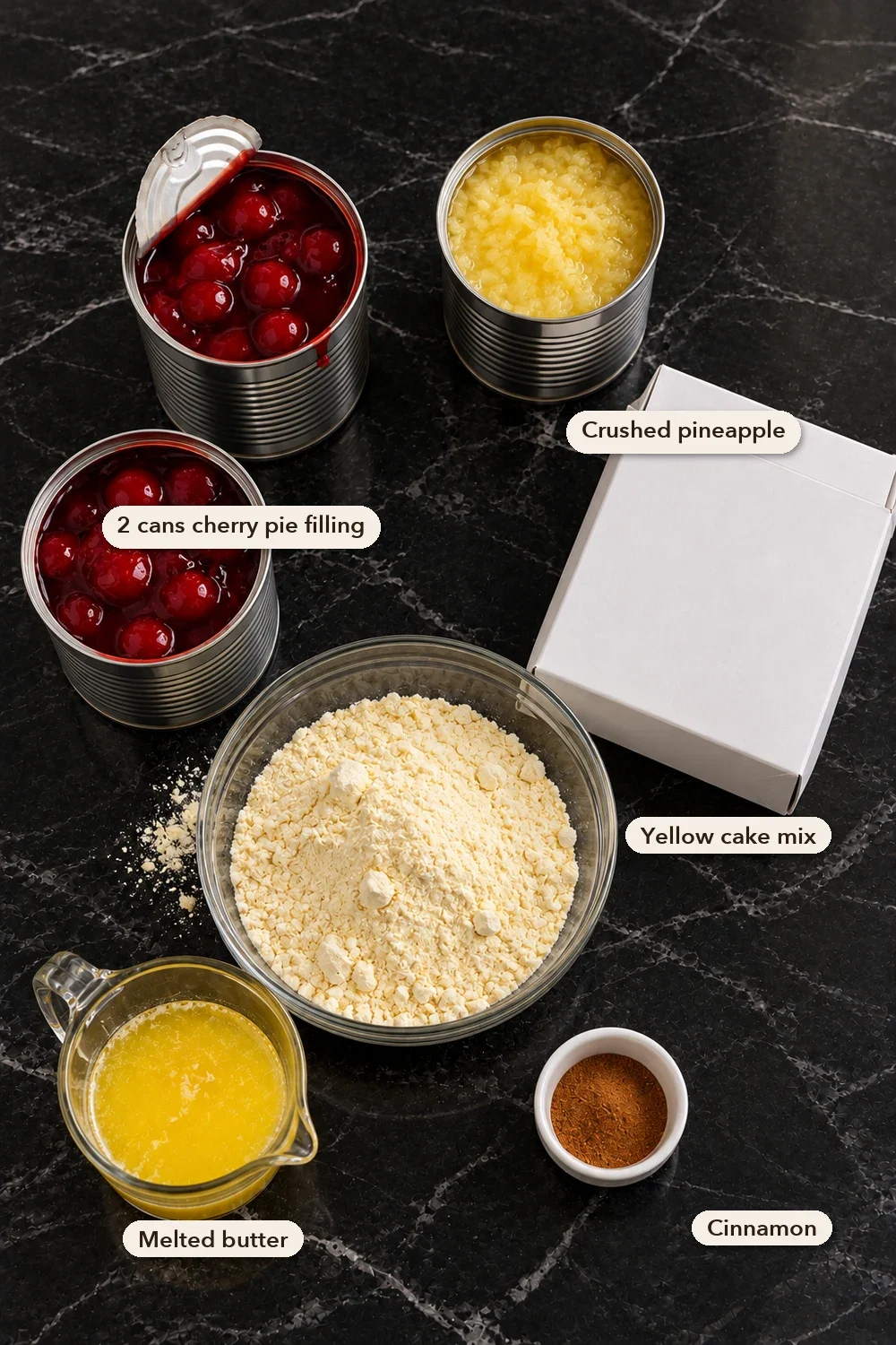 Labeled ingredients on black marble seen from above: two open cans of cherry pie filling, a can of crushed pineapple, a plain box of yellow cake mix beside a glass bowl of the dry mix, a cup of melted butter and a small dish of cinnamon.