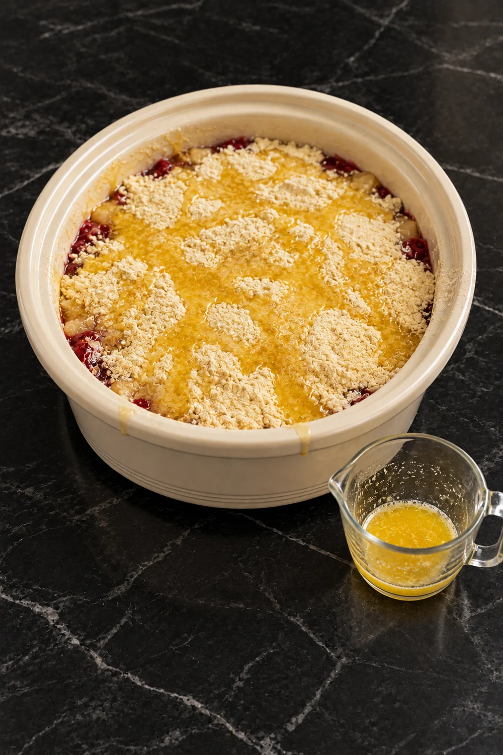 The uncooked layers in a cream slow cooker crock: dry yellow cake mix covering the cherry and pineapple fruit, soaked in streaks and puddles of melted butter, with a small glass measuring cup of butter beside it.