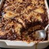 German chocolate dump cake in a white 9x13 baking dish with one corner scooped out, showing a dark chocolate top with golden cream cheese swirls, toasted coconut and pecan halves, and a gooey coconut pecan layer underneath next to a serving spoon.