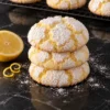 A stack of three thick lemon crinkle cookies with pale yellow cracks under a heavy powdered sugar coat on a black marble counter, a lemon half and zest curls beside them and more cookies on a cooling rack behind.