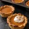 Two mini pumpkin pies on a black stone counter in front of a dark muffin tin of more pies, the front one topped with a dollop of whipped cream and cinnamon, showing flaky pleated crust edges around smooth orange pumpkin filling.