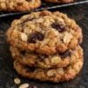 A stack of three oatmeal raisin cookies on a black stone counter with more cookies on a wire rack behind, showing golden edges, whole rolled oats and plump dark raisins on the tops.