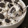 A heaping spoonful of Oreo fluff lifted from a white bowl, showing creamy speckled vanilla cream with chunky Oreo quarters and soft mini marshmallows, with Oreo halves pressed into the top, on a black stone counter.