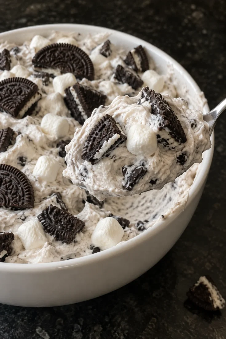 A heaping spoonful of Oreo fluff lifted from a white bowl, showing creamy speckled vanilla cream with chunky Oreo quarters and soft mini marshmallows, with Oreo halves pressed into the top, on a black stone counter.
