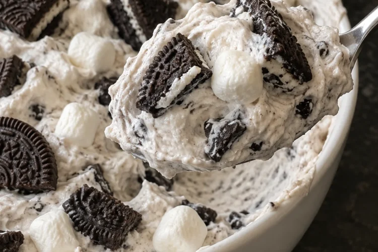 A heaping spoonful of Oreo fluff lifted from a white bowl, showing creamy speckled vanilla cream with chunky Oreo quarters and soft mini marshmallows, with Oreo halves pressed into the top, on a black stone counter.