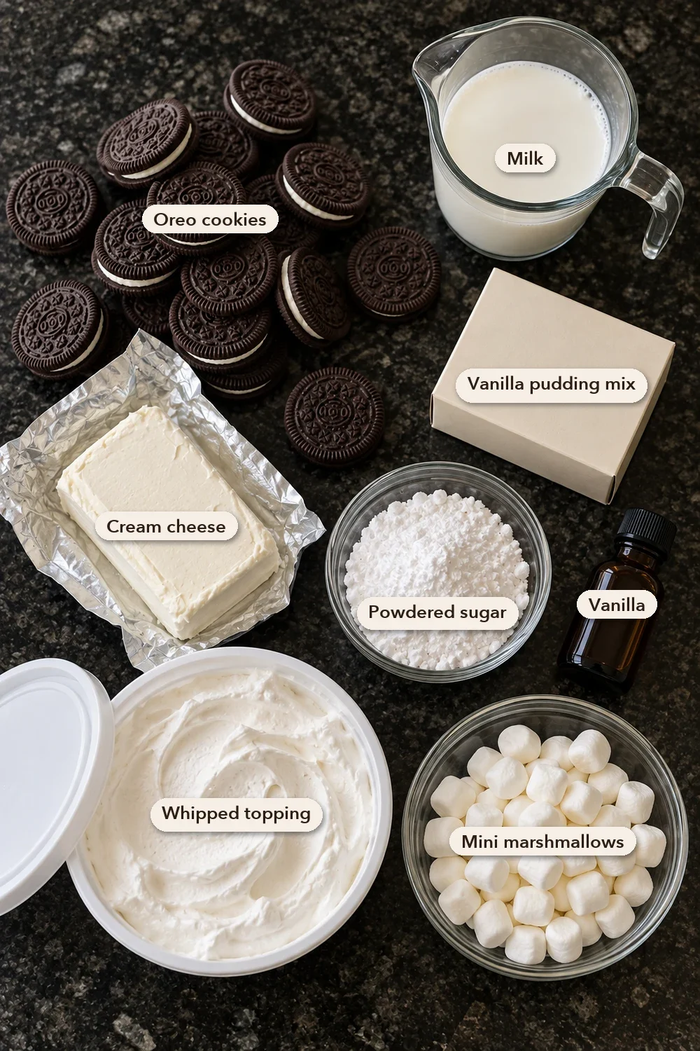 Labeled ingredients for Oreo fluff: Oreo cookies, milk, vanilla pudding mix, cream cheese, powdered sugar, vanilla, whipped topping and mini marshmallows on a black stone counter.