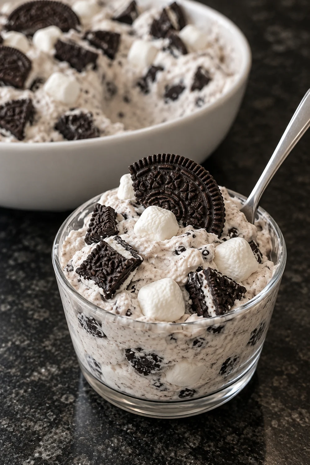 A glass dessert cup of Oreo fluff topped with an Oreo half, marshmallows and a spoon, with the large white bowl of fluff behind it.