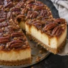 A slice of pecan pie cheesecake lifted from the whole cake on a metal server, showing a graham cracker crust, a tall creamy cheesecake layer and glossy caramel pecans on top, on a black stone counter.